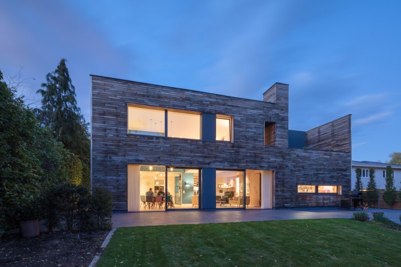 A modern, wooden-cladded house with large windows & patio doors with the inside lights illuminating the surroundings.